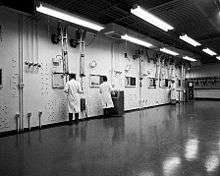 Black and white view of a white wall with six rectangular windows and three men in white coats in front of them, lit by overhead lighting. Long cylindrical rods with handles hang from the ceiling in front of each window.