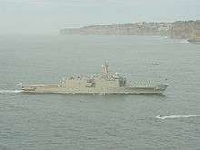 A large grey warship underway. Flags are being flown from the ship's mast, and white-uniformed people line the decks. Brown cliffs line the horizon for the right two-thirds of the image