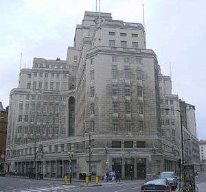 A multi-storey office building in white stone. Two wings containing many small windows meet at right angles. The building is topped by a tower with a flag pole.