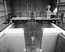 Black and white image of a pool of water with walls partially dividing it. A metal bridge is at the rear of the pool with a long metal assembly hanging from it into the pool. On the bridge at left two men in white lab coats look at a console.