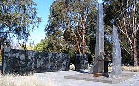 Monument consisting of two upright dark stones in front of a dark stone wall, with trees in the background