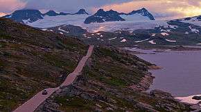 A road through a barren landscape with a lake to the right and mountains in the background