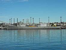 A small warship tied up alongside at a wharf. Her communications and radar masts have been crudely downsized, she carries no weapons, and several large squares have been cut into the ship's hull.