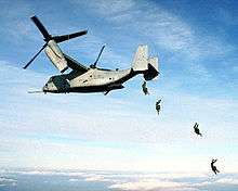 color photo of four parachutists jumping from the open ramp of an MV-22 Osprey in flight