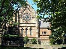 View of the west end of a large church see through trees. There is a rose window in the Decorated Style, and no central door. The entrance porch can be seen projection to the right.