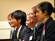 Five high school students in suits and name tags face left. They are seated on the same side of the table and smiling.