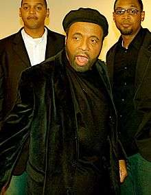 An Afro-American old man, wearing black shirt and black jacket, and with a cap atop his head. He looks a little to the right, and stands with his mouth open. Behind him, two young Afro-American youths can be seen, with the one on right wearing spectacles.