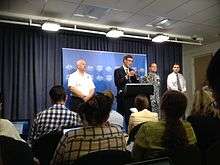 In a small conference room, a seated audience listens to Angus Houston speaking behind a podium and flanked by three other officials.