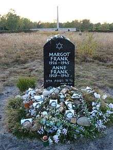 A Memorial for Margot and Anne Frank shows a Star of David and the full names, birthdates, and year of death of each of the sisters, in white lettering on a large black stone. The stone sits alone in a grassy field, and the ground beneath the stone is covered with floral tributes and photographs of Anne Frank