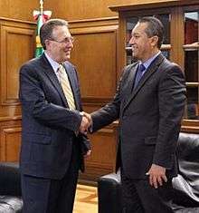 Two men in formal suits shaking hands inside an office.