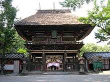A two-storied wooden gate with a large thatched roof and a veranda with handrail on the upper floor.