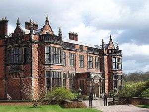 A brick hall seen from a slight angle with bay windows, brick diapering, a porch, shaped gables, and statues standing on the parapets