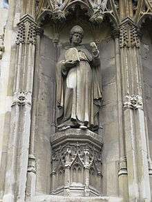 Stone statue of a standing man wearing robes and a mitre, carrying a tablet in one hand and holding his other hand up.