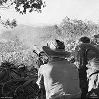 Soldiers firing a machine-gun towards a hillside in the distance. A large number of discarded ammunition belts are visible.