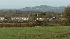 White painted houses with red roofs. In the background is a hill with a tower on it and in the foreground grass and hedgerows