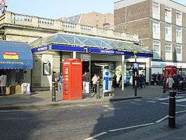 A single storey pale brick building topped with stone railing. Above an entrance is a canopy, around which are rectanglar blue signs reading "BAYSWATER" and "BAYSWATER STATION". People are walking in the street.