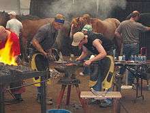 Two men lean over an anvil. One is holding down a red-hot piece of metal with tongs while the other prepares to strike with a large hammer.