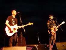 Two men, with an acoustic bass and acoustic guitar, perform on a stage.