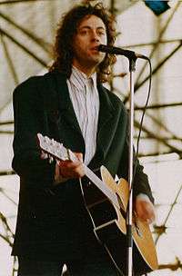 Bob Geldof, a Caucasian man in his mid-thirties, is on stage, singing into a microphone and playing a left-handed acoustic guitar. He wears a white shirt and a dark green jacket.