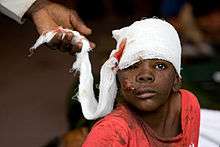 A black child stares through one eye. A hand attends to a bandage which covers his head and that has blood seeping through it. The boy's face is cut and injured. He wears a T-shirt that is sodden and dirty.