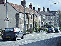 Street scene showing gray stone houses on the left of a road with a few cars.