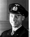 A smiling man wearing a peaked cap and dark military uniform with an Iron Cross displayed at the front of his uniform collar.