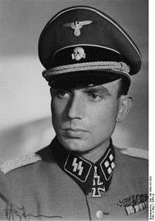 A man wearing a military uniform, peaked cap and a neck order in the shape of a cross. His cap has an emblem in shape of a human skull and crossed bones.