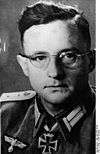 A man with glasses wearing a military uniform with an Iron Cross displayed at the front of his uniform collar.