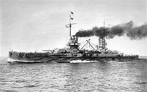 A large gray battleship steams ahead. Dark black smoke pours from its two funnels.