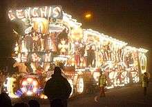 Night time photograph of lorry and trailer illuminated with thousands of light bulbs to make pictures. In the foreground and to the side are pedestrians.