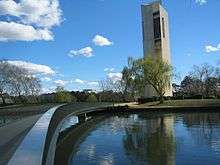 Tall light gray building with irregularly shaped walls and tall narrow windows stretches into the sky before a tree and lake, a bridge crosses the lake to the building.