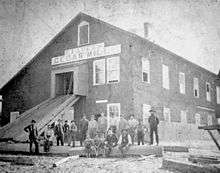 A black and white photograph of a large building featuring a sign that reads "E Faber's Cedar Mill"; More than a dozen white men sit on a large cedar log in the foreground