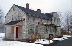 A two-story gray wooden house with light green shutters seen in winter, with snow on the ground. A long wing with two brick chimneys projects towards the left from a higher rear section. At lower right is a black mailbox with "5Ave" on it next to a clear paved driveway