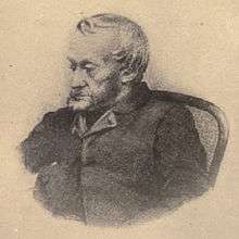 Black-and-white depiction from the waist up of an old, bearded man dressed in a dark jacket, sat in a chair and looking towards the left.