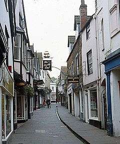 Street scene. Buildings lining narrow lane with central water gully, pedestrians, and hanging baskets.