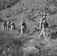 Three unarmed soldiers being led down a hill by two soldiers armed with rifles, one in the front the other in the rear.