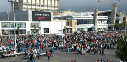 Several thousand people stand in lineups while volunteers serve food. A large shopping mall stands in the background.