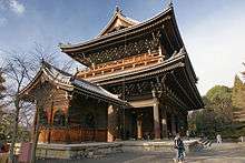 A large two-storied wooden gate with a veranda on the upper story and a hip-and-gable style roof. A small wooden building with gabled roof is placed next to it.