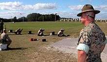 color photo of a rifle range, with recruits firing rifles at distant targets while a Warrant Officer observes