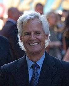 A headshot of a smiling man with long white hair and a black suit