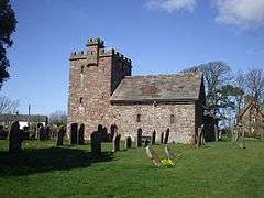 A stone church seen from the south, with a large squat battlemented west tower and a small body, both which contain very small windows