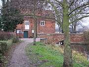 A view of the mill building from the footpath on the north bank.  The pool and the bridge across the race are visible to the right, but the red brick face of the building dominates. In the extreme foreground on the left is a bare-branched tree, its trunk lightly flecked with lichen. In the middle distance on the left is a holly, dark green and dense.