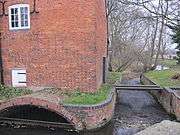 Close view of the south side of the building.  A small eliptical brick arch leads the water into the enclosed millrace, and there is an overflow weir to the right of the building. Trees are visible on the riverbank beyond.  The water is constrained in a red brick channel with a bulnose (curved) corner.  Two scroll shaped iron 'plates' are on the upper wall, terminals for tie-rods that pass through the building. This is a winter view, and the state of the trees makes this stark. There is a small and unturbulent flow of water through the overflow weir, because the level is accurately controlled by the main weir in the old lock chamber. A precarious plank bridge, with no handrails, crosses the overflow race to reach the lockside on the right.