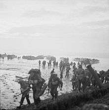 Men in the foreground burdened with equipment, behind them are mud or sand flats and in the distance landing craft