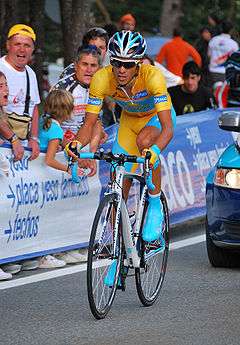 A man in yellow clothes and blue shoes, riding a bicycle, followed by a car. People are watching him from behind a fence.