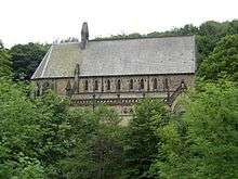 A stone church seen through foliage with an apse to the left, a nave and chancel with a clerestory, and a bellcote towards the left