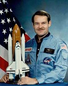 man in flight suit holding Space Shuttle mode, American flag in background