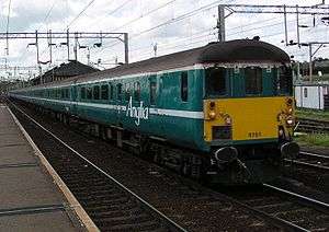 Stationary train with a yellow front and turquoise livery with a white stripe down its length. Electrified wires are above the train and track.