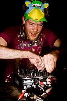 A white man with red hair and a slight beard manipulating the controls of a piece of music mixing equipment. He has headphones around his neck and a mask resembling a multi-coloured monkey pushed up onto the top of his head.