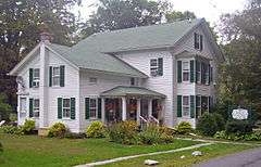 A white house with green shutters, a light green roof and bright flowers out front